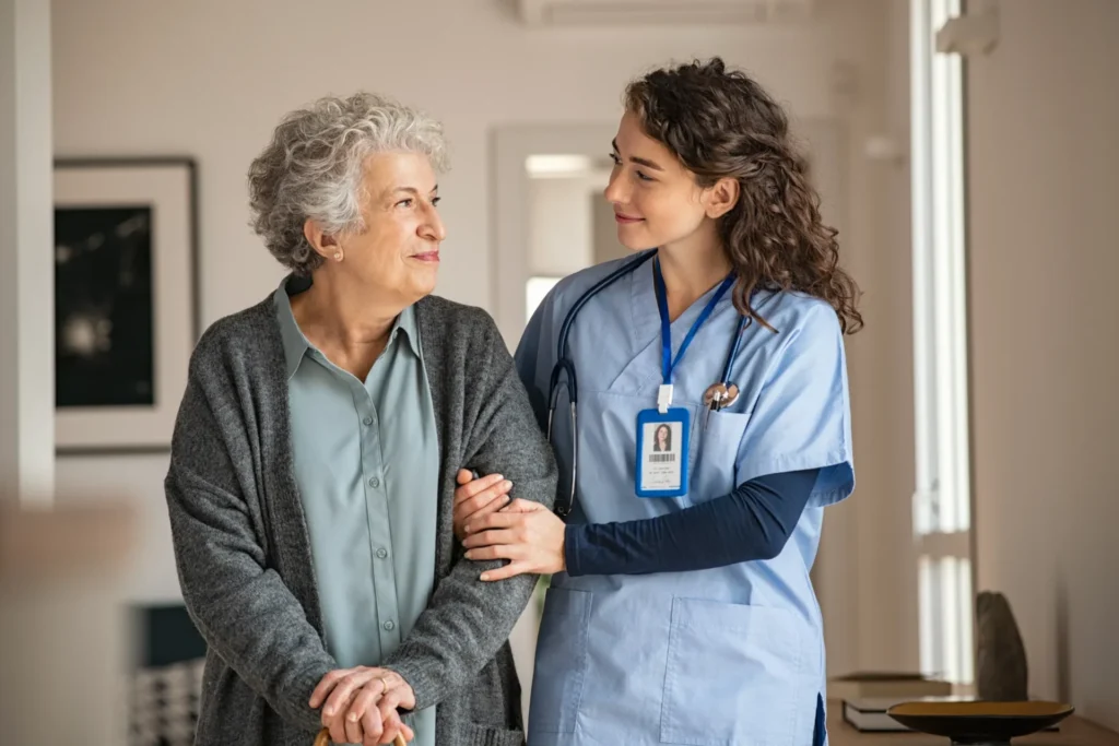 A young nurse warmly assists an elderly woman as they walk together. They share a kind and reassuring smile in a well-lit home setting.